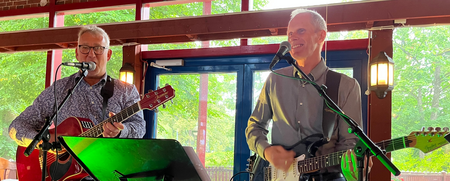 Två män spelar gitarr och sjunger på en scen inomhus med stora fönster i bakgrunden.