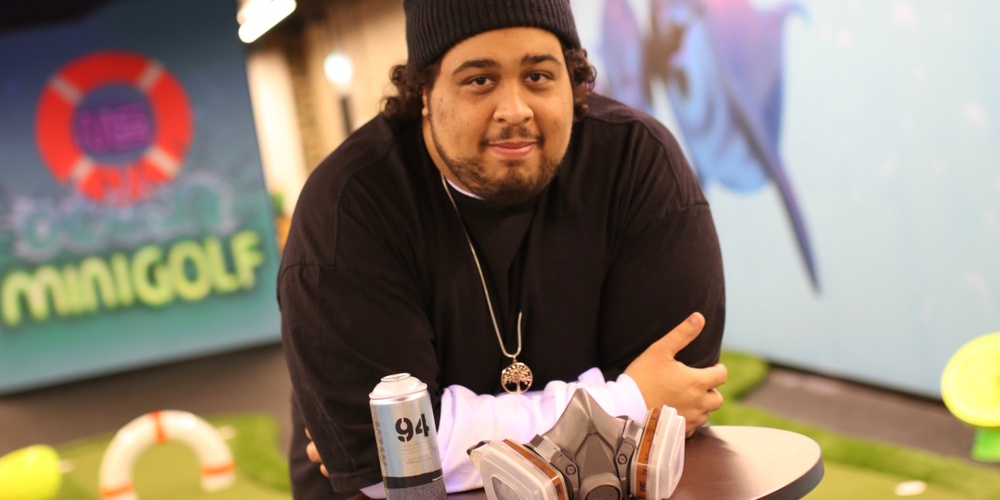 Man med mössa lutar sig mot ett bord med sprayburk och ansiktsmask i en inomhus minigolfmiljö.