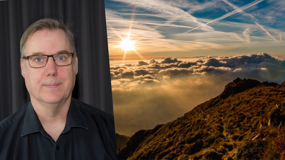 En person i svart skjorta står framför ett mörkt draperi, bredvid ett landskap med solnedgång över moln och berg.