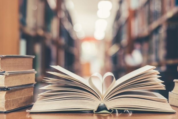 En uppslagen bok ligger på ett bord i ett bibliotek, omgiven av staplade böcker.