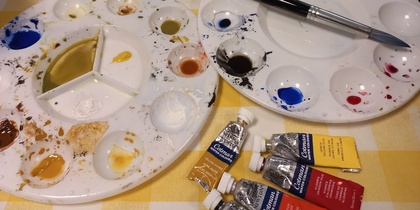 Två målarpaletter med färg, pensel, tuber med akrylfärg och två målningar på ett bord med gulrutig duk.