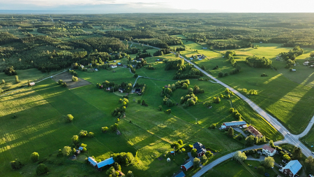 Flygfoto över ett grönt landskap med åkrar, skog, vägar och utspridda hus i kvällssol.