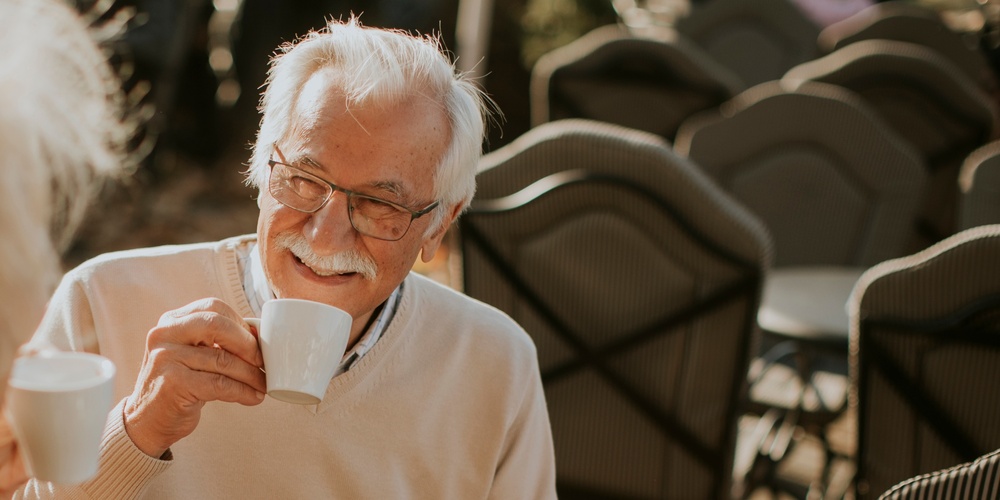Två äldre personer sitter utomhus vid ett runt bord och dricker kaffe tillsammans.