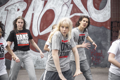 En grupp unga personer dansar energiskt utomhus framför en graffitivägg, alla bär tröjor med texten 'Dans Studio'.
