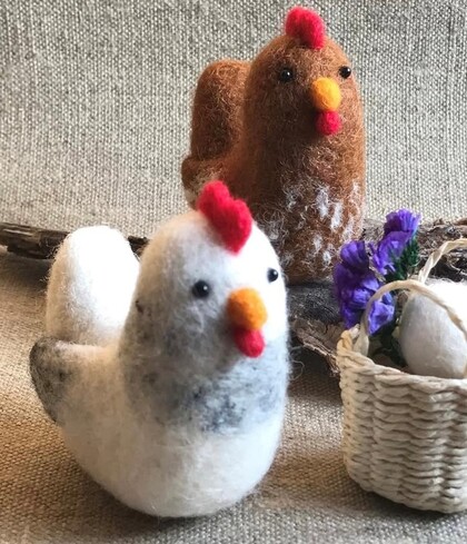 Två tovade hönor, en vit och en brun, står på ett tygunderlag bredvid en liten korg med lila blommor.