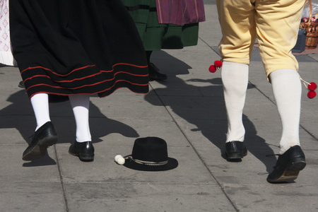 Två personer i traditionella folkdräkter dansar på en stenlagd yta, en svart hatt ligger på marken.