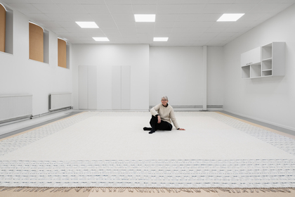 En person sitter på ett stort, ljust matta i ett minimalistiskt, vitt rum med högt i tak.