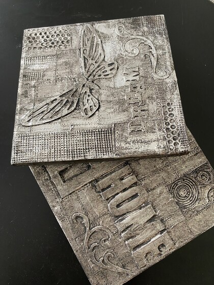 Två dekorativa fyrkantiga plattor med reliefmönster, text och fjärilsmotiv i svart och silver.