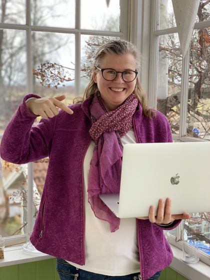 Leende kvinna med glasögon och lila jacka håller en bärbar dator och pekar på den vid ett fönster.