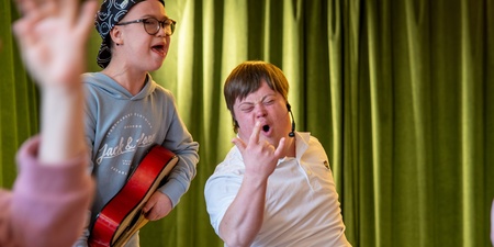 Två unga personer, en med gitarr, sjunger och gör rockposer framför gröna gardiner.