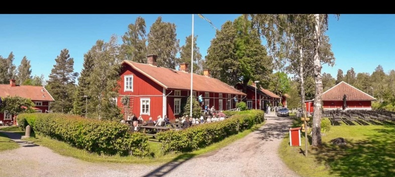 Röda trähus med vita knutar omgivna av grönska, uteservering och grusväg under klarblå himmel.