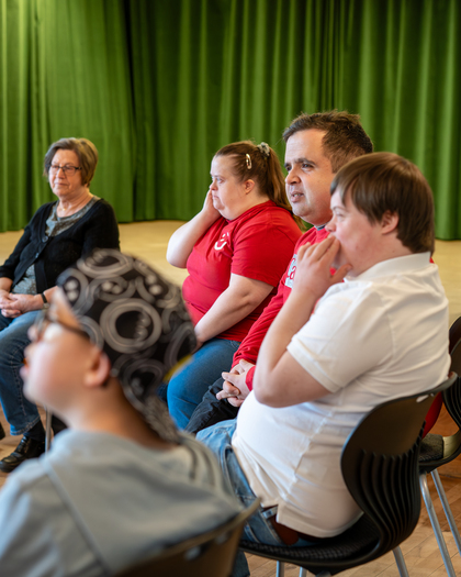 En grupp personer sitter i en cirkel i ett rum med grön gardin i bakgrunden.