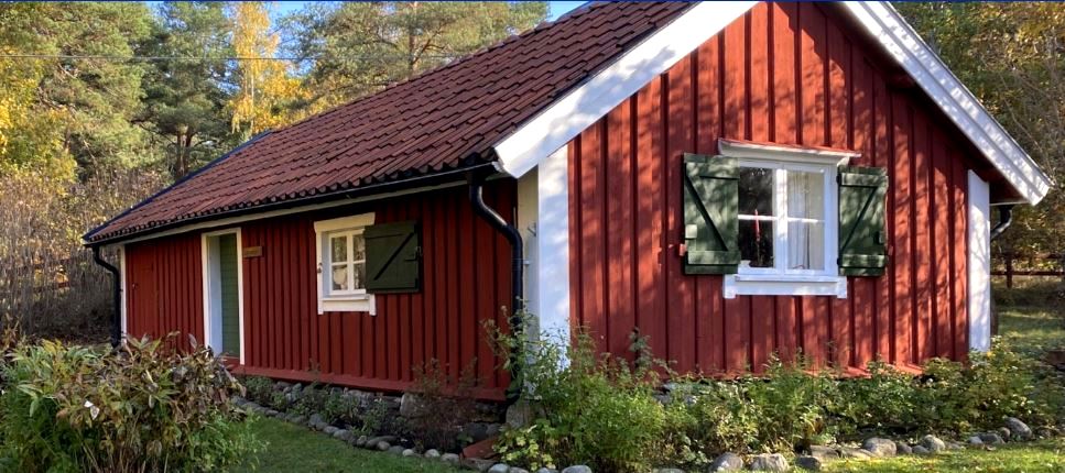 Röd stuga med vita knutar och gröna fönsterluckor i en grönskande trädgård.