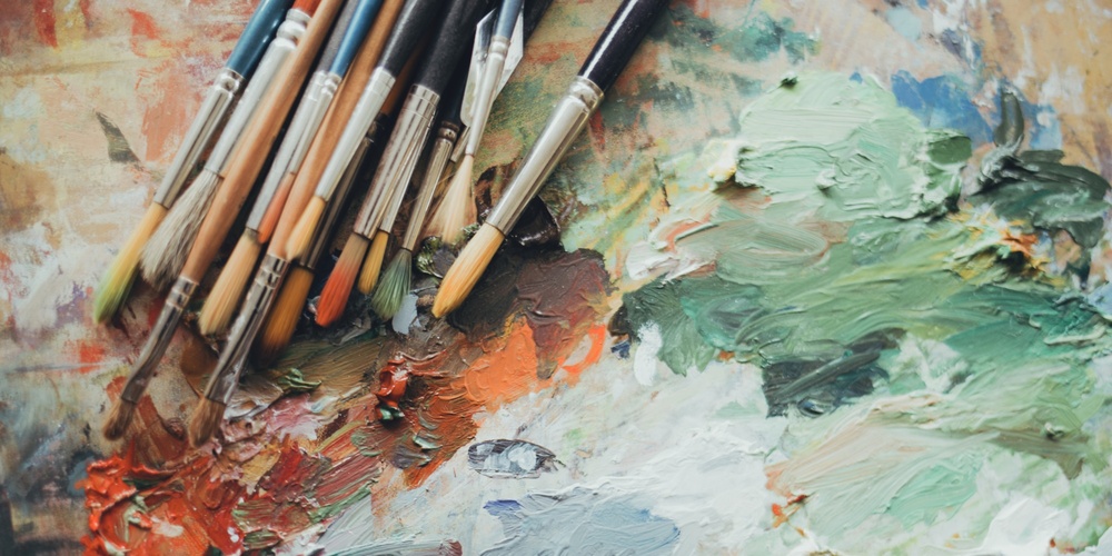 Flera penslar ligger på en färgpalett med blandade oljefärger i grönt, vitt och orange.