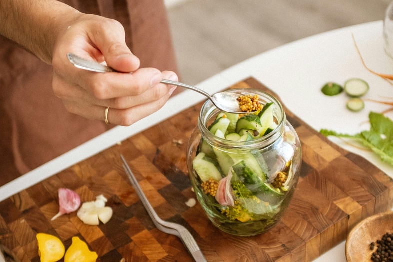 En person lägger grönsaker i en glasburk på en skärbräda, omgiven av ingredienser för inläggning.
