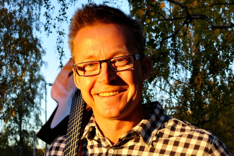 Leende man med glasögon och gitarr på axeln står utomhus i kvällssol, klädd i rutig skjorta.