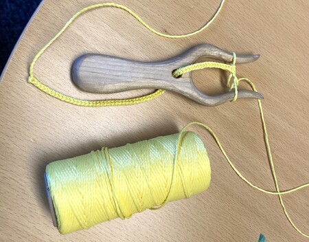 En trådgaffel i trä med gul tråd och en rulle gul tråd på ett träbord.