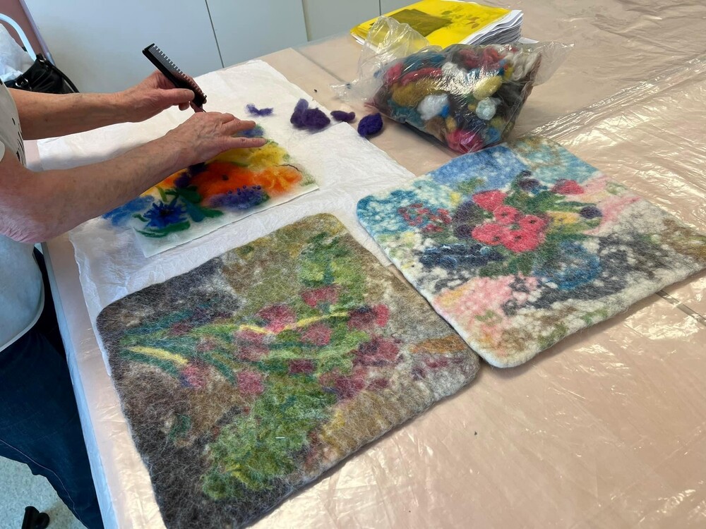 En person arbetar med tovning av färgglada ullbitar på en vit handduk, två färdiga tovade konstverk ligger bredvid.