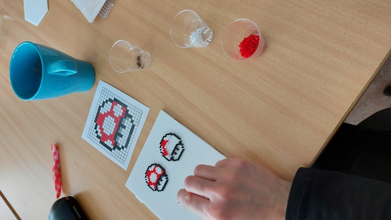 En person gör pärlplattor med motiv av svampar från Super Mario på ett bord med muggar och små plastkoppar.