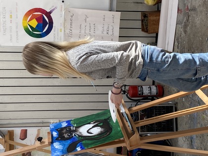 Person målar en katt på duk vid ett staffli i en konstsal med färgcirkel och instruktioner på väggen.