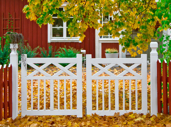 Vit trägrind framför ett rött hus, omgiven av höstlöv och träd med gula blad.