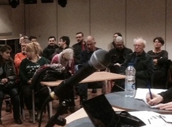 Ett möte eller seminarium med människor som sitter i ett rum, några vid ett bord med mikrofon och papper.