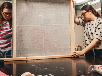 Två personer arbetar tillsammans med en stor träram i en verkstad med textilmaterial.