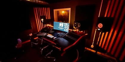 Ett mörkt inspelningsstudio med orange belysning, dator, högtalare och ljudutrustning på ett skrivbord.