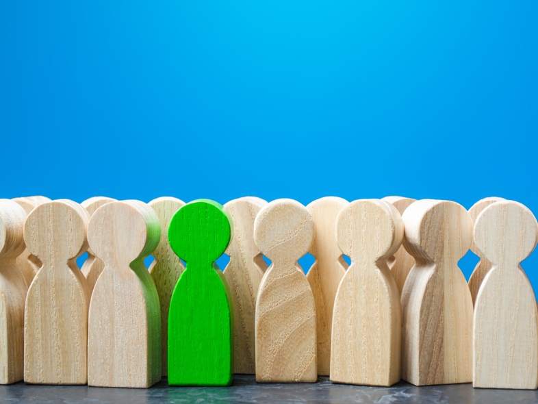 Träfigurer står i rad, en av dem är grön medan resten är träfärgade, mot en blå bakgrund.