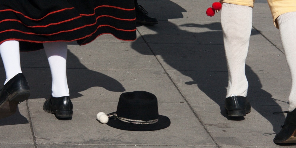 Två personer i traditionella folkdräkter dansar på en stenlagd yta, en svart hatt ligger på marken.