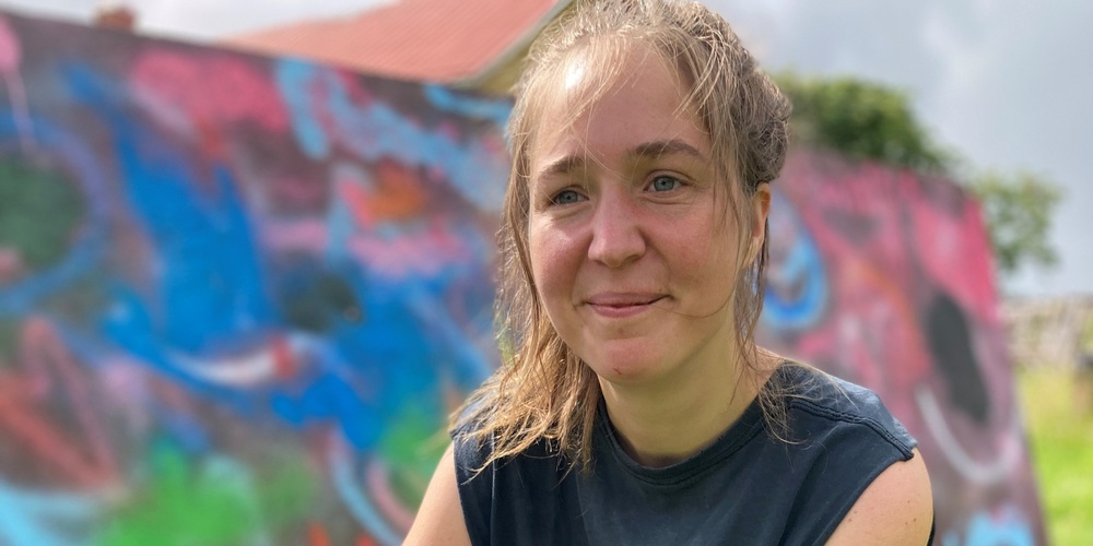 En kvinna sitter på huk på gräs framför en färgglad graffitivägg utomhus.