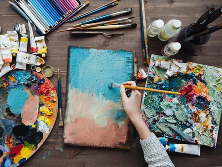 En person målar med pensel på en duk omgiven av färgtuber, penslar och paletter på ett träbord.