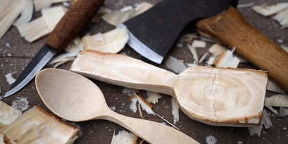 En träsked, en yxa och en kniv ligger på ett bord omgivet av träspån och en halvfärdig träsked.