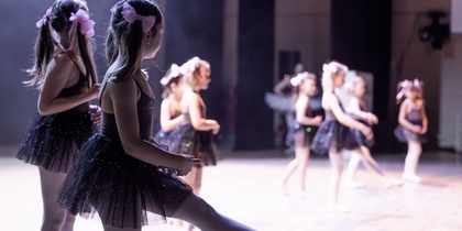 Flera unga flickor i svarta balettklänningar dansar på en scen med ljus bakgrund.