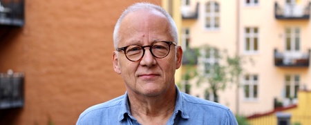 Äldre man med glasögon och blå skjorta står utomhus framför färgglada byggnader.