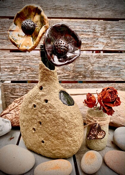 Två konstgjorda blommor i en rustik vas av papier mache, bredvid en mindre vas med torkade rosor och stenar på ett träbord.