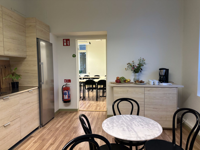 Ljust kök med runda bord, svarta stolar, frukt, kaffebryggare och utsikt mot ett klassrum.