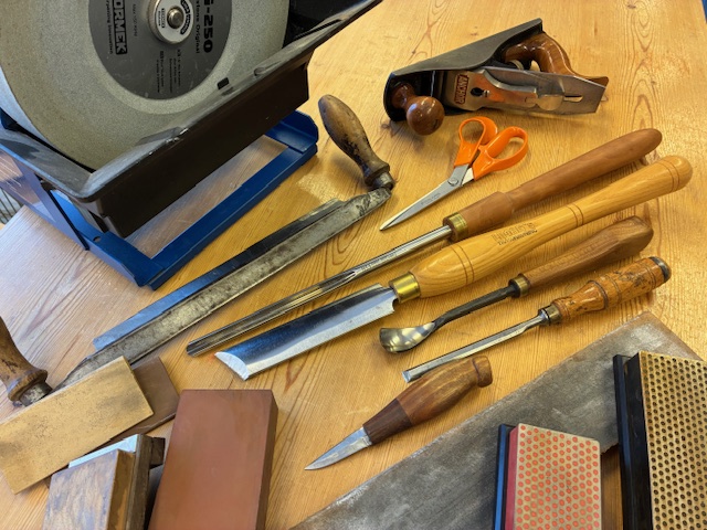 Ett bord med olika snickeriverktyg, slipsten, hyvel, sax, filar, stämjärn och slipstenar.