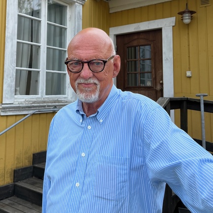 Äldre man med glasögon och blå randig skjorta står utomhus framför ett gult trähus med vita fönsterkarmar.
