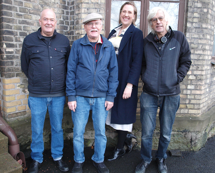 Fyra personer utan instrument står utomhus framför en tegelvägg, tre män och en kvinna.