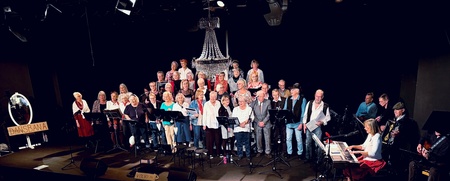 Kör och musiker uppträder på scen inför publik i en teaterlokal med mikrofoner och instrument.