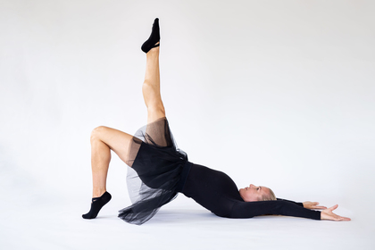 En person i svart dansdräkt gör en bro med ett ben rakt upp i luften på vit bakgrund.