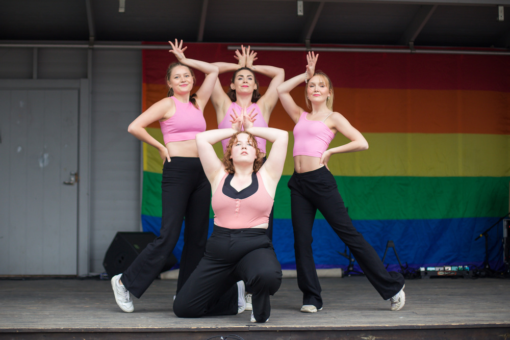 Fyra unga kvinnor i rosa toppar och svarta byxor dansar på en scen framför en regnbågsflagga.