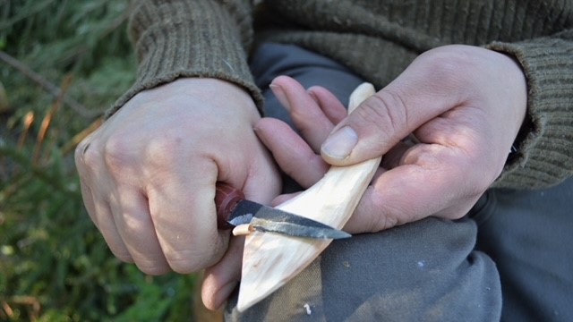 En person täljer en träbit med en kniv, händerna syns tydligt i närbild.