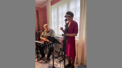 Två personer musicerar, en spelar gitarr och en sjunger vid mikrofon i ett rum med röda väggar och vita gardiner.
