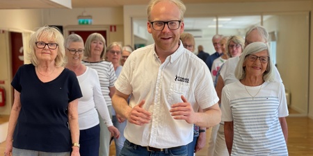 En grupp äldre personer och en yngre man står tillsammans inomhus, leende och avslappnade.