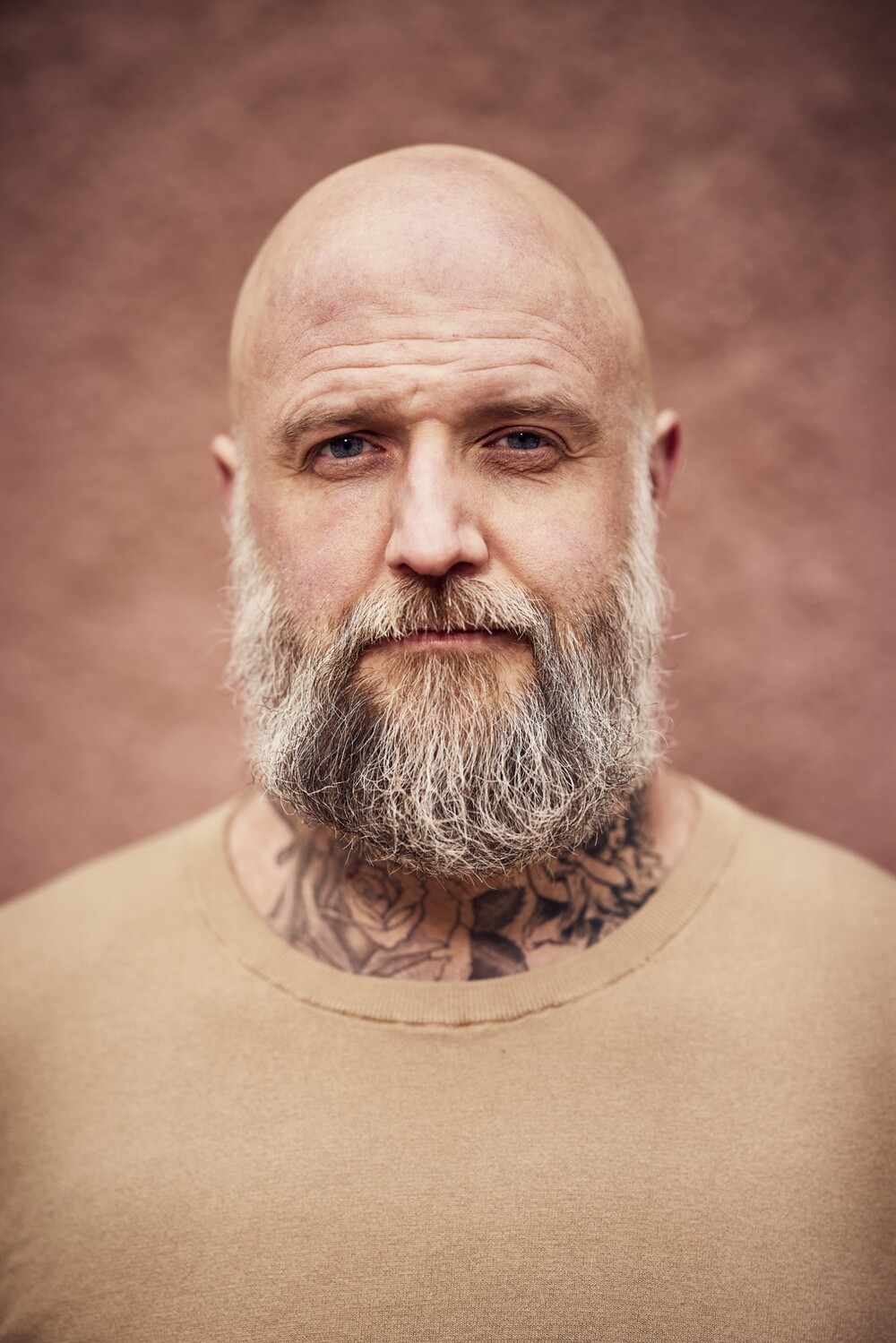 Skäggig, skallig man med tatueringar på halsen, beige tröja och neutral bakgrund.