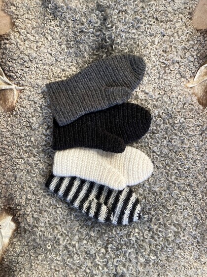 Fyra nålbundna vantar i grått, svart, vitt och svartvitt randigt ligger på en lockig grå ullpläd.