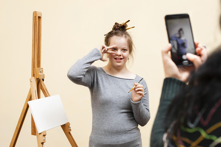 Ung kvinna med penslar i håret poserar framför ett staffli medan någon tar ett foto med mobiltelefon.