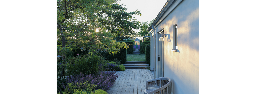 Trädgård med trädäck, grönska och modern husfasad i kvällssol.